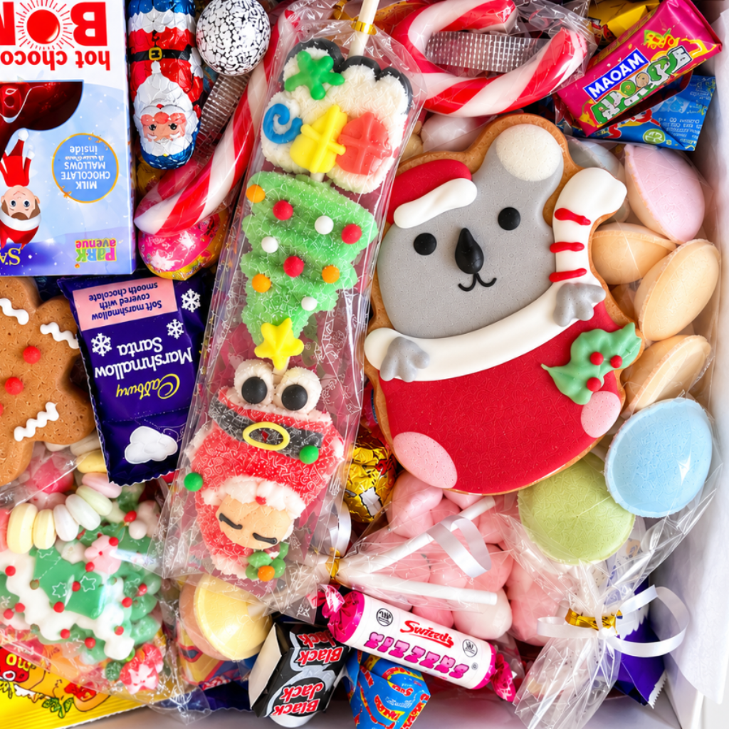A Christmas-themed candy and cookie gift box filled with assorted sweets, decorated cookies, and packaged treats. Visible items include Santa-themed cookies, candy canes, wrapped chocolates, a gingerbread person cookie, and a Santa-themed hot chocolate bomb in a box.