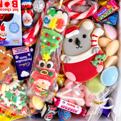 A Christmas-themed candy and cookie gift box filled with assorted sweets, decorated cookies, and packaged treats. Visible items include Santa-themed cookies, candy canes, wrapped chocolates, a gingerbread person cookie, and a Santa-themed hot chocolate bomb in a box.